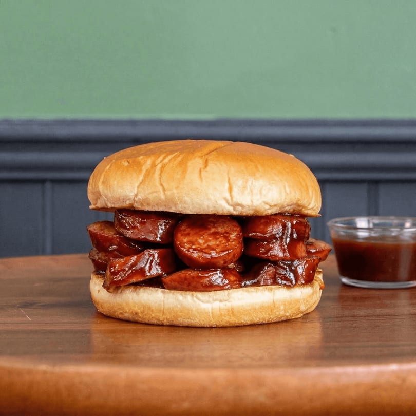 Shelby's Craft BBQ sausage sandwich with barbecue sauce on a wooden table.
