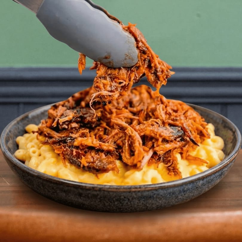Tongs adding pulled pork to a bowl of creamy mac and cheese from Shelby's Craft BBQ.