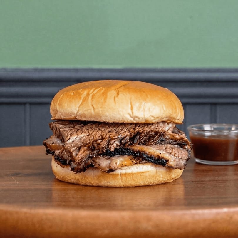 Shelby's Craft BBQ brisket sandwich on a bun with a side of barbecue sauce.